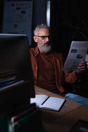 serious bearded businessman looking at paper with graphs while sitting near empty notebook at night in officeの写真素材