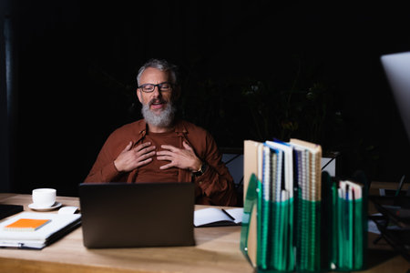 satisfied businessman touching chest and looking at laptop near coffee cup and notebooks in dark officeの写真素材