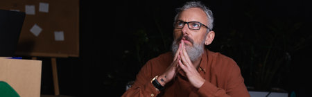 mature businessman in eyeglasses sitting with praying hands and looking up in office at night,...