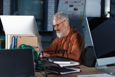 Mature businessman in casual clothes and eyeglasses using computer near notebooks in officeの写真素材