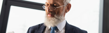 Positive businessman in eyeglasses looking at blurred smartphone in office, bannerの写真素材
