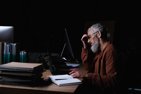 side view of thoughtful and tired businessman sitting near laptop and blank notebook at night in...