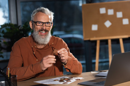 bearded businessman in eyeglasses smiling at camera near bitcoins and laptop at workplaceの写真素材
