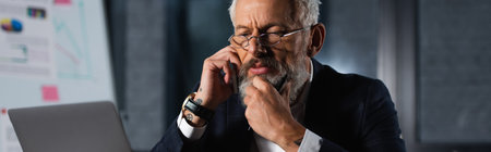 Bearded businessman in eyeglasses talking on smartphone near laptop in office in evening, banner