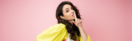 coquettish woman in yellow blouse looking at camera and showing hush sign isolated on pink, banner