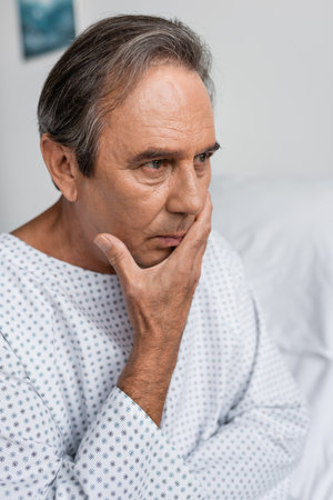Disappointed elderly man in patient gown looking away in hospital ward