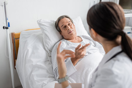 Senior patient talking to blurred doctor with paper folder in hospital wardの写真素材
