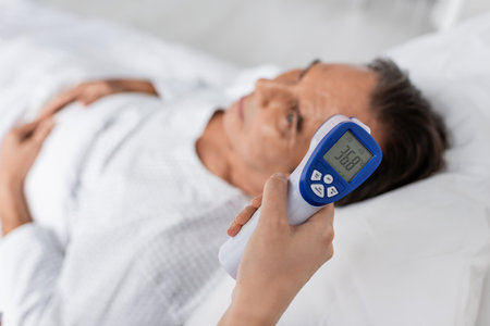 Doctor holding pyrometer near blurred elderly patient in clinic