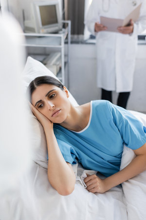 Displeased woman in patient gown lying on bed near blurred doctor in hospital ward
