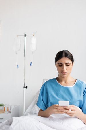 brunette patient using cellphone while sitting on bed in hospitalの写真素材
