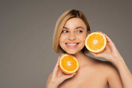 cheerful woman with bare shoulders holding orange halves isolated on greyの写真素材