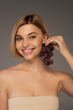 joyful young woman with bare shoulders holding ripe grapes near ear isolated on greyの写真素材