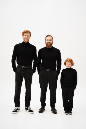 full length of cheerful bearded men with red haired kid in black clothes standing with hands in pockets on light grey backgroundの写真素材
