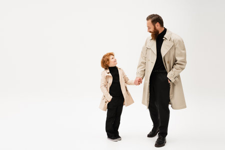 happy redhead kid and bearded man in trench coats holding hands and looking at each other on light grey backgroundの写真素材