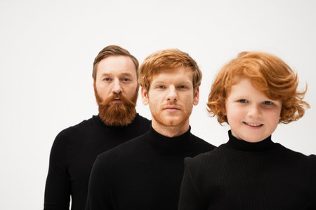 bearded man in black turtleneck looking at camera near redhead son and happy grandson isolated on greyの写真素材