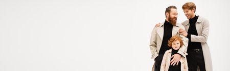 young man in trench coat hugging bearded father near redhead son looking at camera isolated on grey, bannerの写真素材