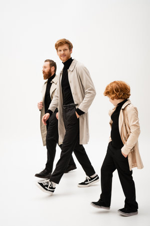full length of trendy bearded men and redhead boy in trench coats walking on light grey backgroundの写真素材