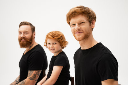 smiling man with redhead son and bearded tattooed father sitting and looking at camera isolated on greyの写真素材