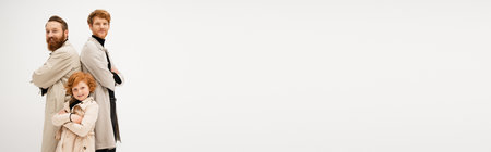 cheerful kid in trench coat posing with crossed arms near redhead dad and grandpa standing back to back isolated on grey, bannerの写真素材