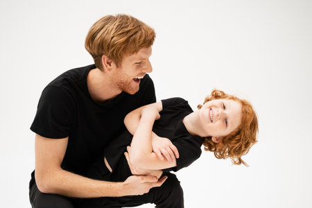 excited man in black t-shirt playing with happy redhead son crossing arms isolated on greyの写真素材