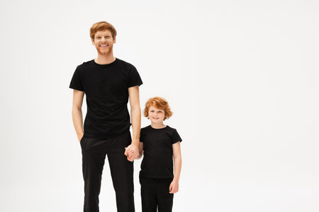 joyful redhead father and son in black t-shirts and trousers holding hands and smiling at camera isolated on greyの写真素材