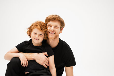 pleased man in black t-shirt embracing red haired kid and looking at camera isolated on greyの写真素材