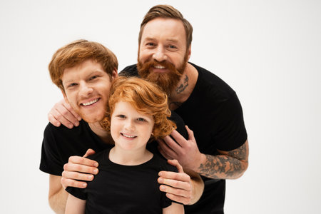 family portrait of redhead men and cheerful boy in black t-shirts hugging and smiling at camera...