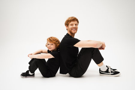 full length of happy red haired dad and son in black t-shirts and pants sitting back to back on light grey backgroundの写真素材