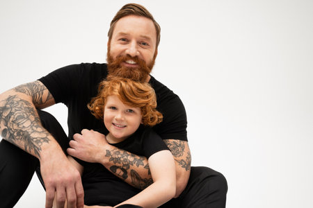 cheerful tattooed man in black t-shirt hugging redhead grandson while sitting isolated on greyの写真素材