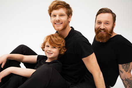 joyful red haired kid with father and bearded grandpa in black t-shirts looking at camera isolated on greyの写真素材