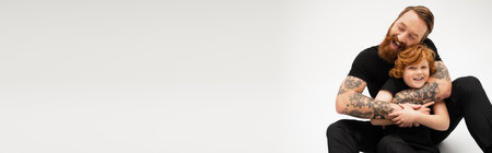 excited tattooed man with closed eyes hugging red haired grandson while sitting on grey background, bannerの写真素材