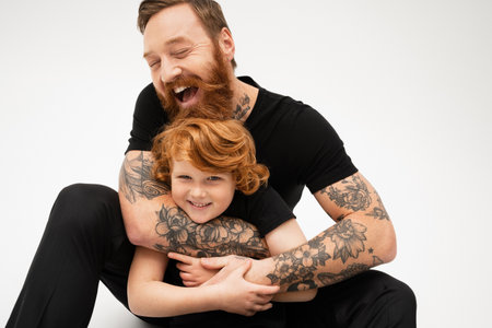 cheerful tattooed man with closed eyes hugging happy redhead grandson on grey backgroundの写真素材