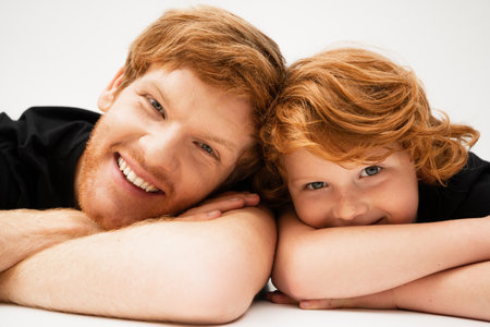 family portrait of redhead father and son lying with crossed arms and smiling at camera on light grey backgroundの写真素材