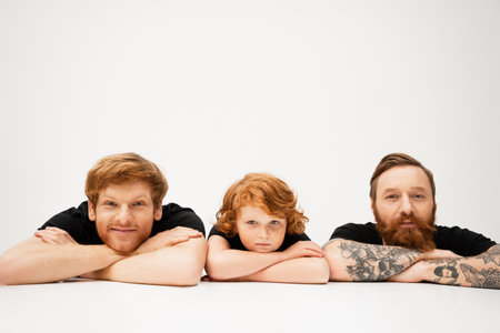 cheerful man with redhead son and bearded tattooed father lying with crossed arms on light grey backgroundの写真素材