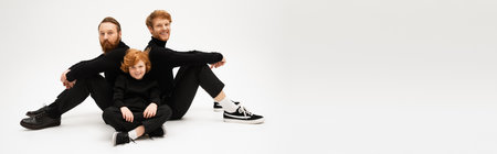 happy red haired boy sitting with crossed legs near bearded grandpa and father on light grey background, bannerの写真素材