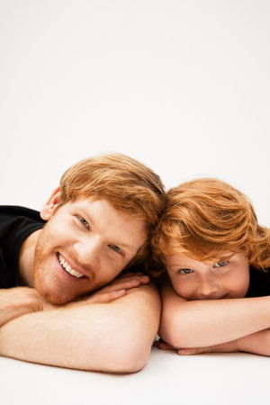 young and cheerful man with red haired son lying with crossed arms and looking at camera on light grey backgroundの写真素材