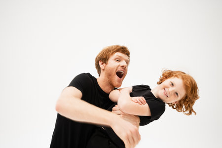 excited man playing with redhead son crossing arms and smiling at camera isolated on greyの写真素材