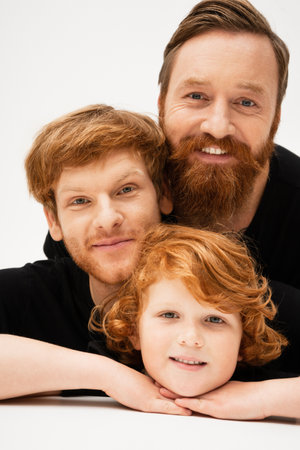 family portrait of redhead kid with dad and bearded grandfather smiling at camera on light grey backgroundの写真素材