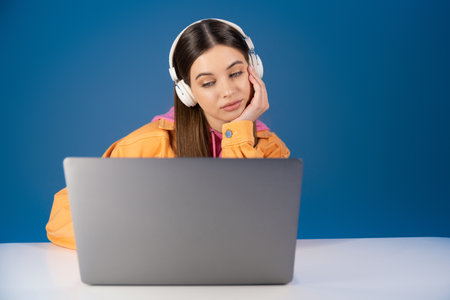 Brunette teenager in headphones looking at laptop on table isolated on blueの写真素材
