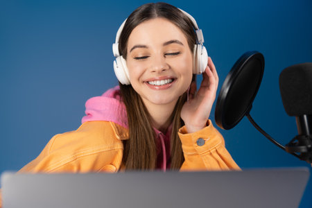 Pleased teenager in headphones looking at laptop near microphone isolated on blueの写真素材