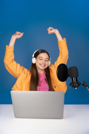 Excited teenager in headphones using laptop and studio microphone and sitting with raised hands isolated on blueの写真素材