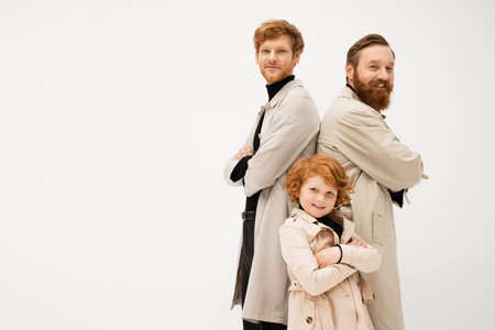 joyful bearded man in trench coats standing back to back with crossed arms near redhead boy isolated on greyの写真素材