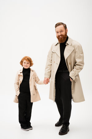 full length of cheerful red haired boy with grandfather holding hands while posing in trench coats on light grey backgroundの写真素材