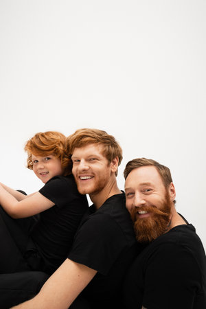 happy bearded man with redhead son and grandson in black t-shirts smiling at camera isolated on greyの写真素材