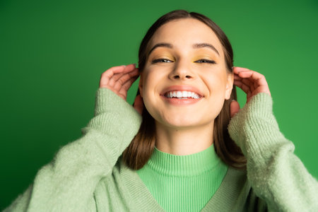 Cheerful teenager in cozy jumper adjusting hair isolated on greenの写真素材