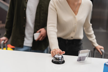 partial view of woman ringing service bell on reception near blurred boyfriend with smartphone in...