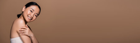 Cheerful asian woman in top touching arm and looking at camera isolated on brown, banner