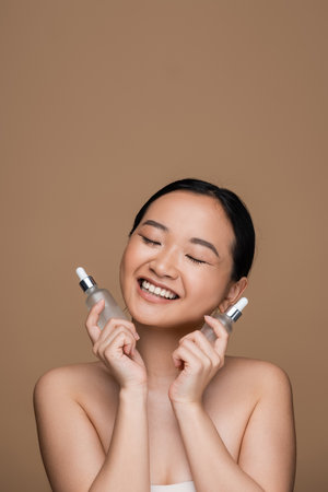 Pleased asian woman holding cosmetic serums isolated on brown