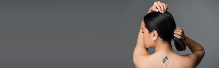 Side view of young tattooed asian woman touching ponytail isolated on grey, banner