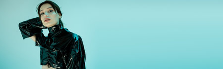 young woman in black latex clothing looking at camera isolated on blue, banner
